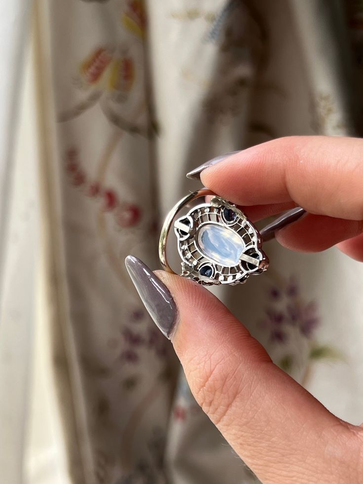 “Art Deco” was an era marked by extravagance and innovation. The Art Deco style itself was an eclectic mix of heavy geometric shapes, with curved ornamental elements. The resulting silhouettes were often monolithic but elegant, with applied decorative elements. This Moonstone and Sapphire Edwardian Conversion Ring is a wonderful example of this iconic Art Deco style. Crafted in 20 carat white gold, this ring immediately captures the gaze with its dazzling array of miniature pearls and sapphire stones haloing a truly luminescent cabochon moonstone. What makes this treasure particularly special is that it is a conversion piece; the beautiful moonstone center was once part of an antique brooch. Now, with the use of a 14 carat gold replica shank, it has been transformed into the glorious ring Luxury Silver Moonstone Ring, Luxury Silver Oval Moonstone Ring, Elegant Multi-stone Moonstone Ring As Gift, White Gold Polished Moonstone Open Ring, White Gold Moonstone Open Ring With Polished Finish, Luxury Silver Moonstone Ring For Formal Occasions, Luxury Oval Jewelry With Ring Detail, Luxury Oval Ring With Detailing, Art Deco Multi-stone Oval Sapphire Ring