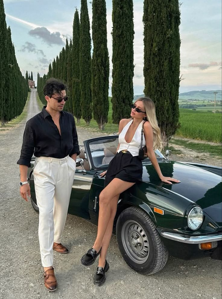 a man and woman sitting on the hood of a green car in front of trees