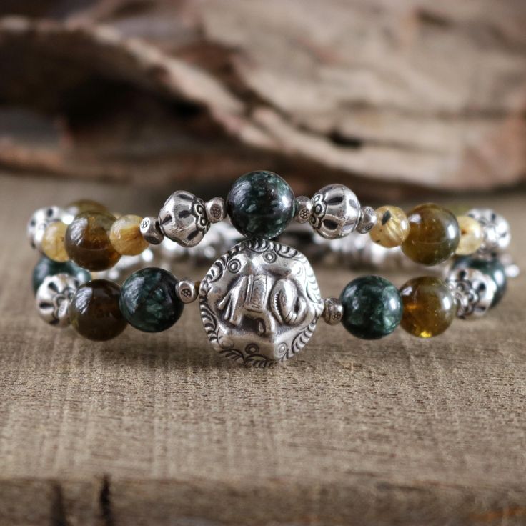 * Add a touch of elegance and cultural charm to your jewelry collection * This handmade bracelet is adorned with intricate Hill Tribe silver charms, adding an ethnic and rustic feel. It is made from Seraphinite beads (10.5 mm), Tsavorite green Garnet beads (10.3 mm), and golden Rutile Quartz beads (6.8 mm), which are beautiful, high-grade natural stones carefully selected to match the color tone. Paired perfectly with a prominent Hill Tribe silver elephant bead and an elephant S-clasp. It also features 925 sterling silver and Hill Tribe fine silver components, renowned for their high purity ranging from 95-99%, surpassing the standard 92.5% sterling silver (certification of silver purity can be viewed on the shop's About page), expertly strung together with a strong and durable waxed cord, Beaded Elephant, Elephant Jewelry, Silver Elephant, Elephant Bracelet, Silver Elephants, About Page, Hill Tribe Silver, Green Garnet, Rutile Quartz