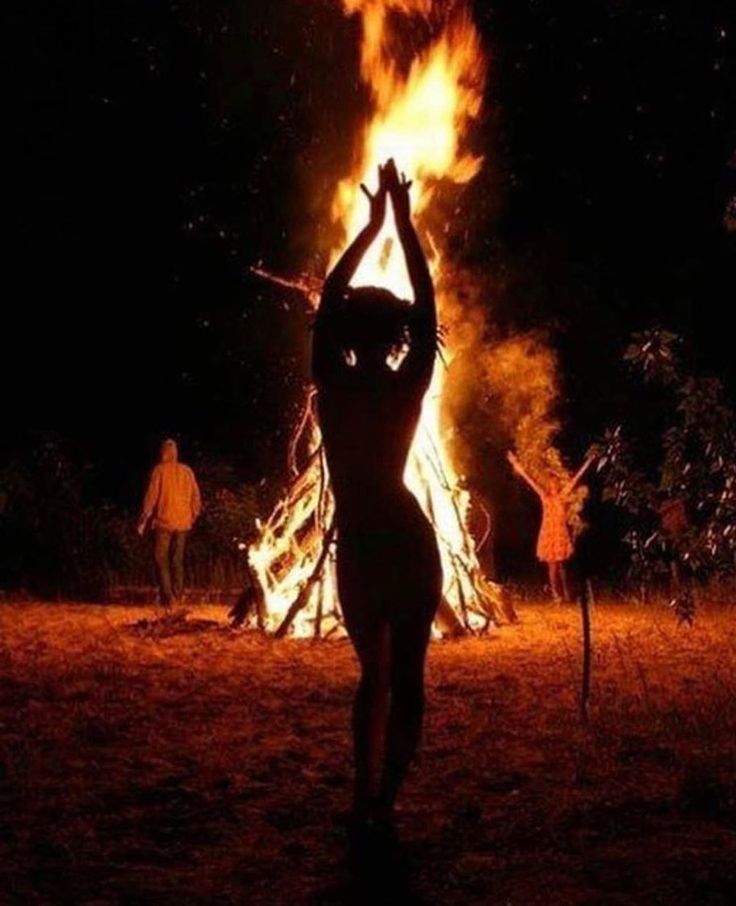 a woman standing in front of a fire with her arms up and hands raised above her head