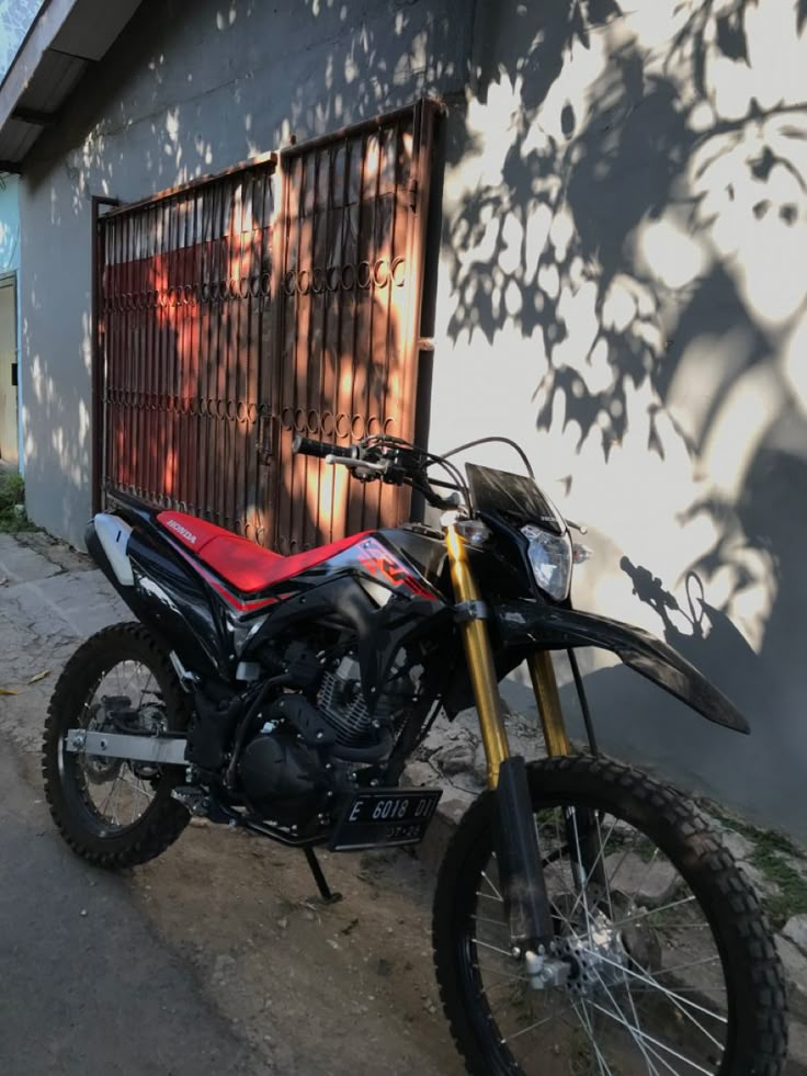 a red and black motorcycle parked in front of a building next to a tree with its shadow on the wall