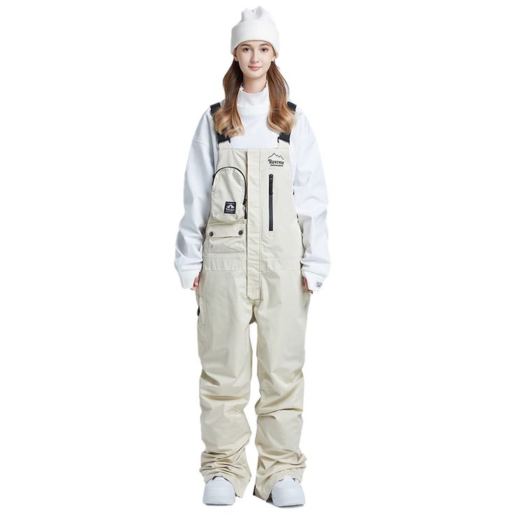 a woman standing in front of a white background wearing overalls and a beanie