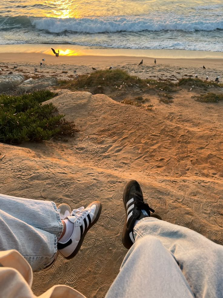 two people sitting on the beach with their feet in the sand