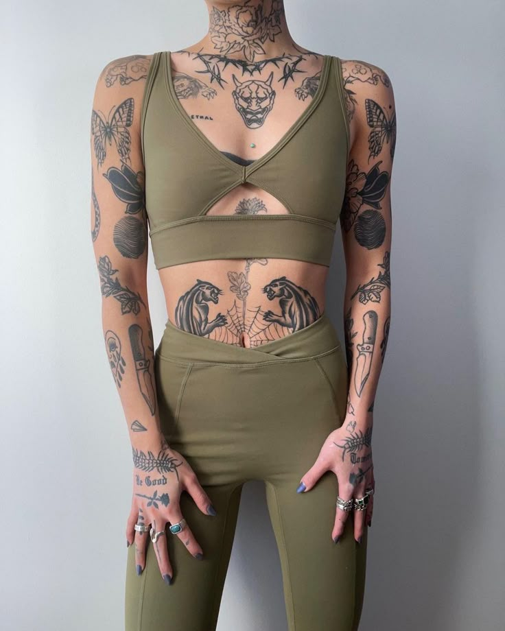 a woman with tattoos on her arms and chest wearing green leggings, standing in front of a white wall