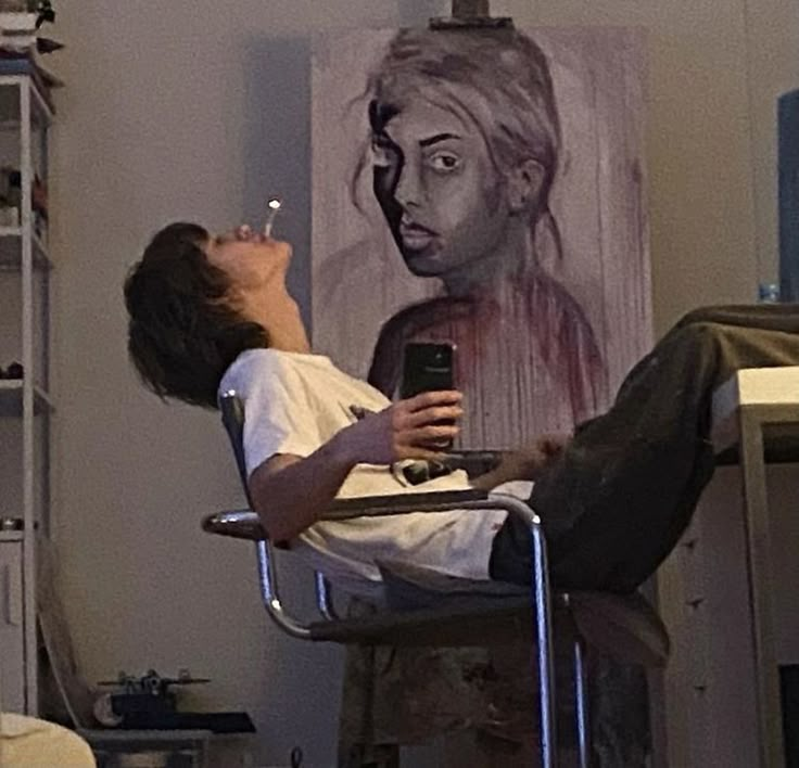 a man sitting in a chair holding a cell phone up to his face while looking at a painting