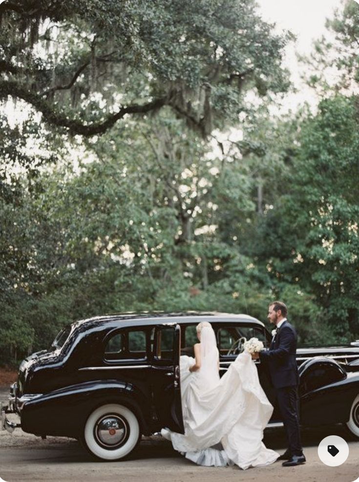 a bride and groom standing in front of an old black car with trees behind them