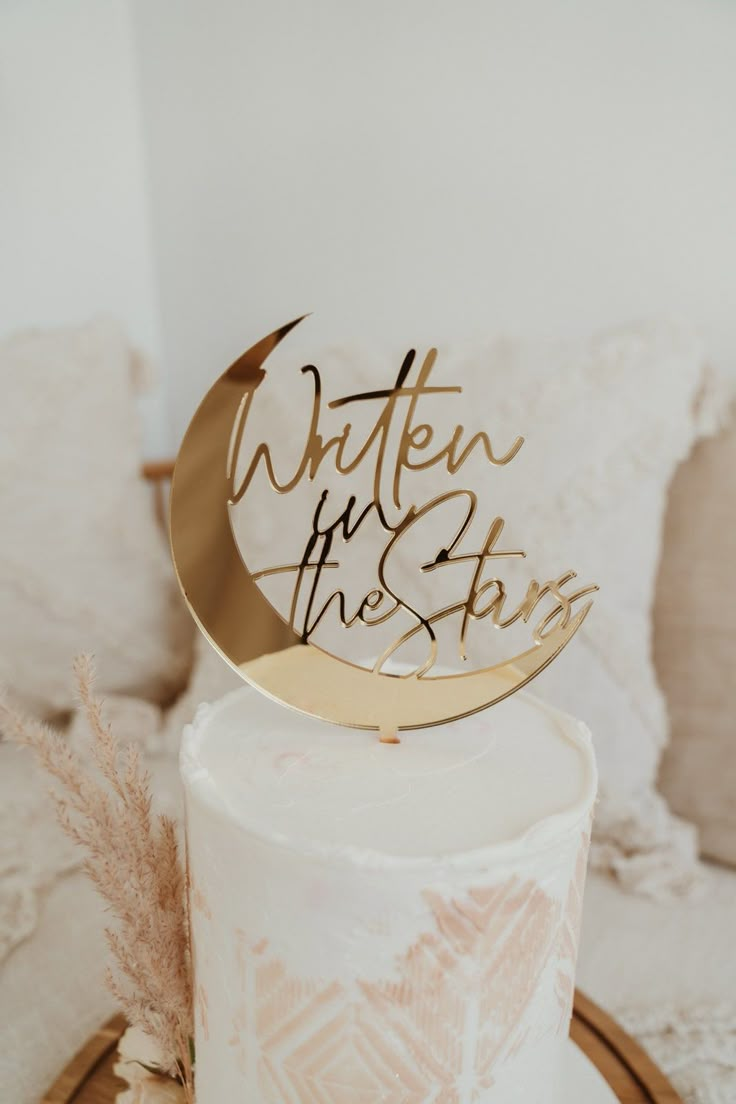 a white cake with gold lettering on top and a round wooden sign that says when in the sky