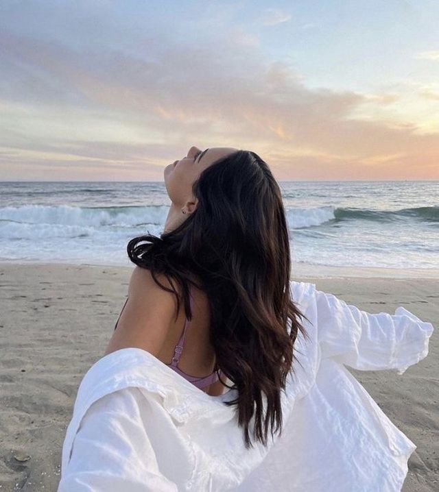 a woman sitting on top of a sandy beach next to the ocean with her arms outstretched