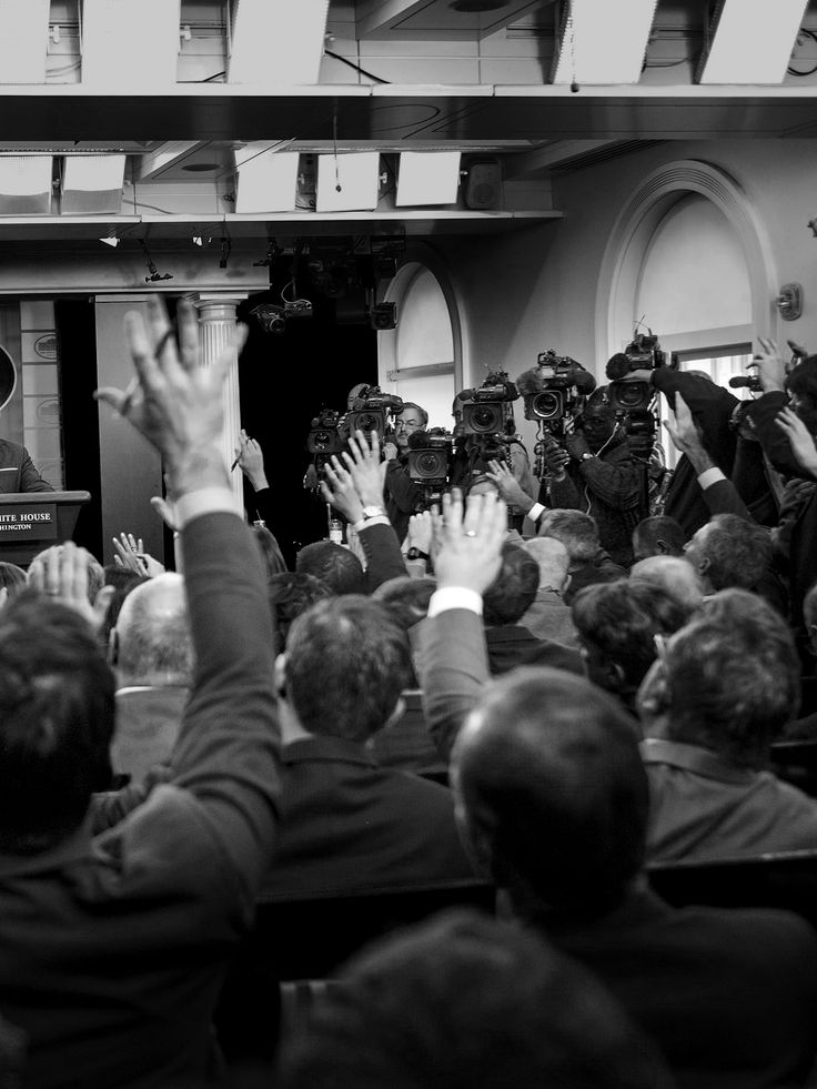 black and white photograph of people raising their hands in front of a podium with cameras around them