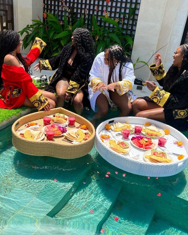 four women sitting on the edge of a swimming pool with plates of food in front of them