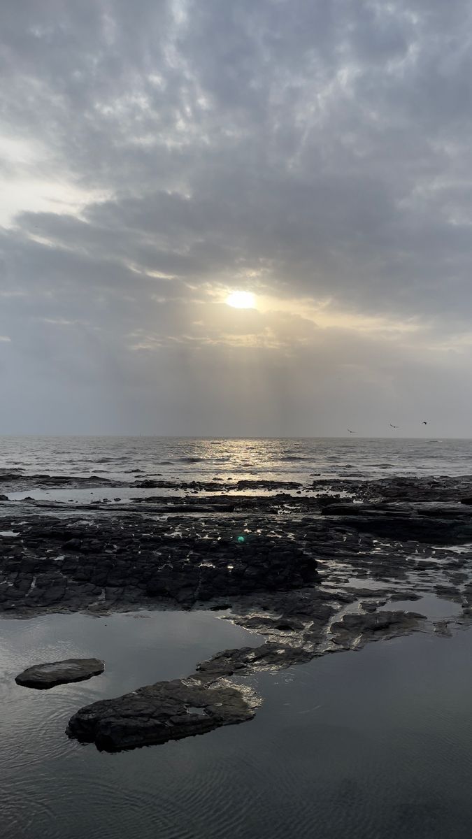 the sun is shining through clouds over the water and rocks on the beach at low tide