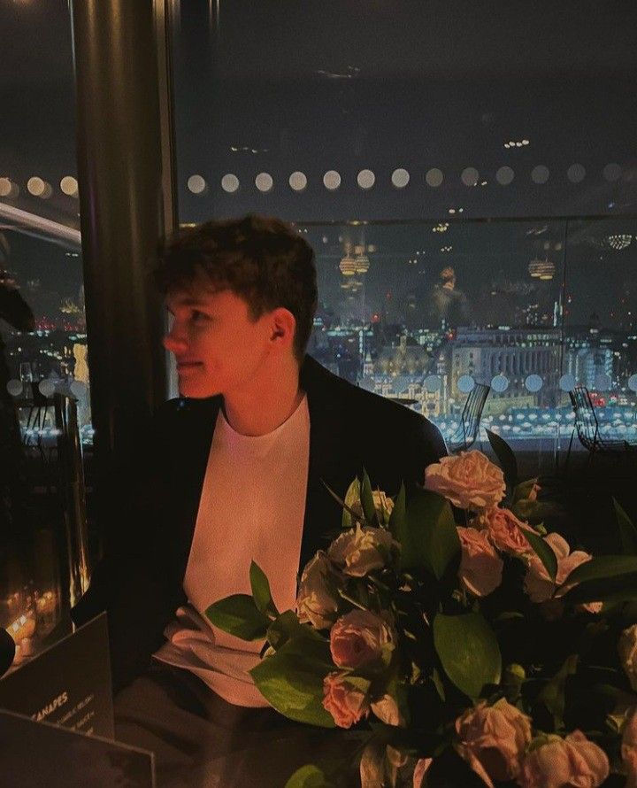 a man sitting at a table with flowers in front of him and the city lights behind him