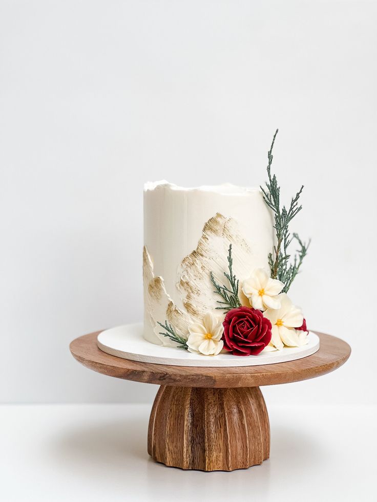 a white cake decorated with flowers and greenery on a wooden stand in front of a white wall