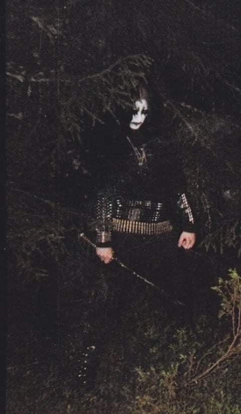 a man with white face paint holding a guitar in the woods at night, wearing a black outfit