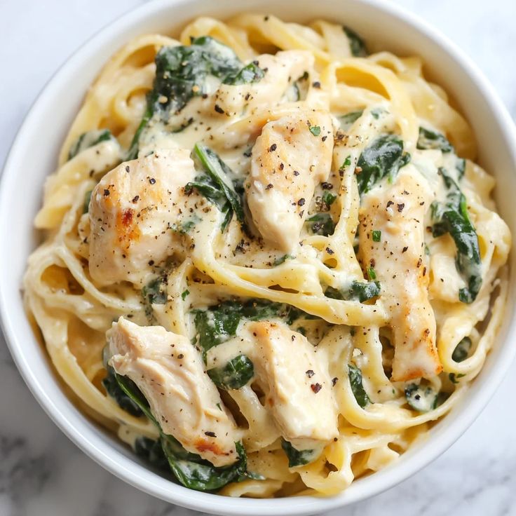 pasta with chicken and spinach in a white bowl on a marble countertop top