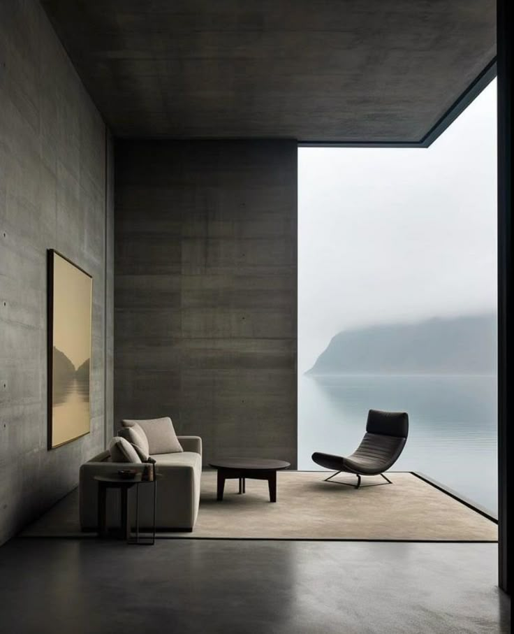 an empty room with a chair, couch and table next to the window looking out on water