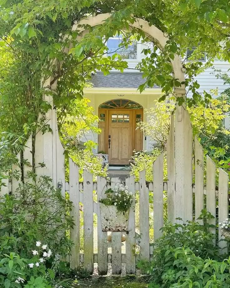 a white picket fence surrounded by greenery and flowers in front of a small house