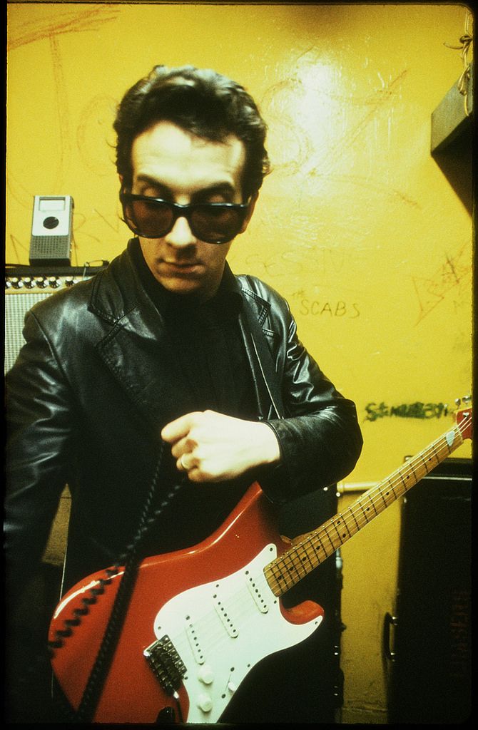 a man wearing sunglasses and holding a red guitar in his right hand while standing next to a yellow wall