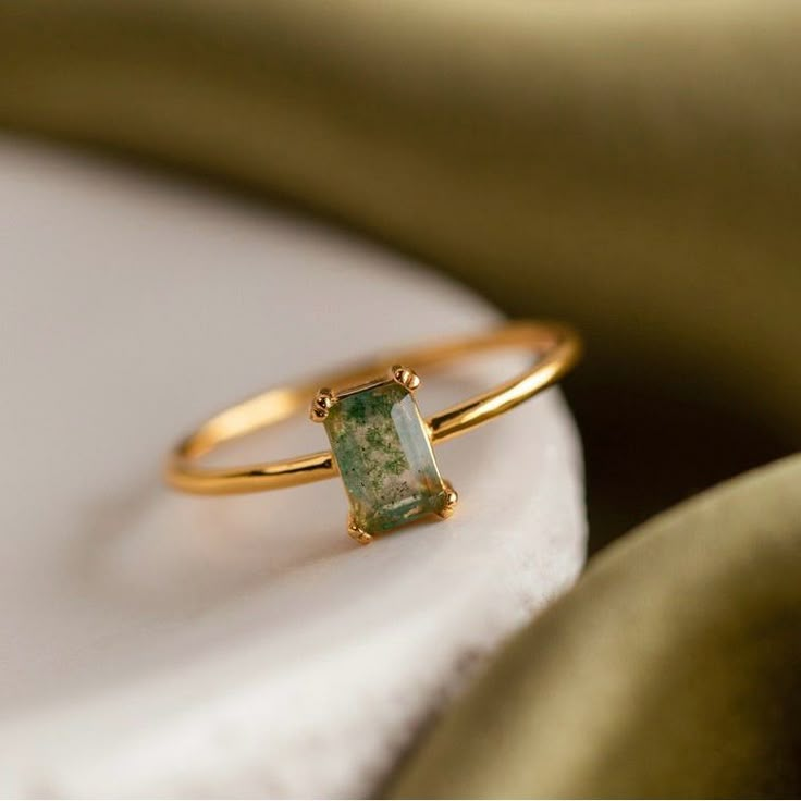 a gold ring with a green stone on it sitting on a white plate next to a golden cloth