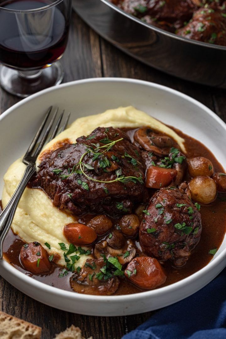 a white plate topped with meat and potatoes covered in gravy next to a glass of wine