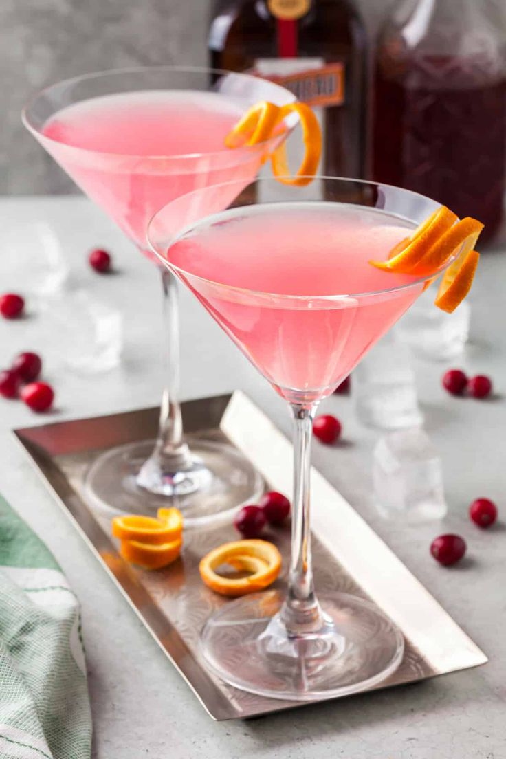 two glasses filled with pink lemonade and garnished with orange peels on a silver tray