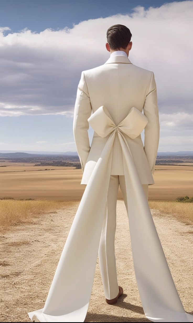 a man in a white suit and bow tie standing on a dirt road with his back to the camera