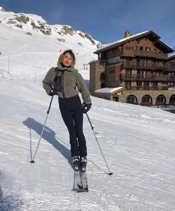 a woman standing on skis in the snow near a building with a mountain behind her