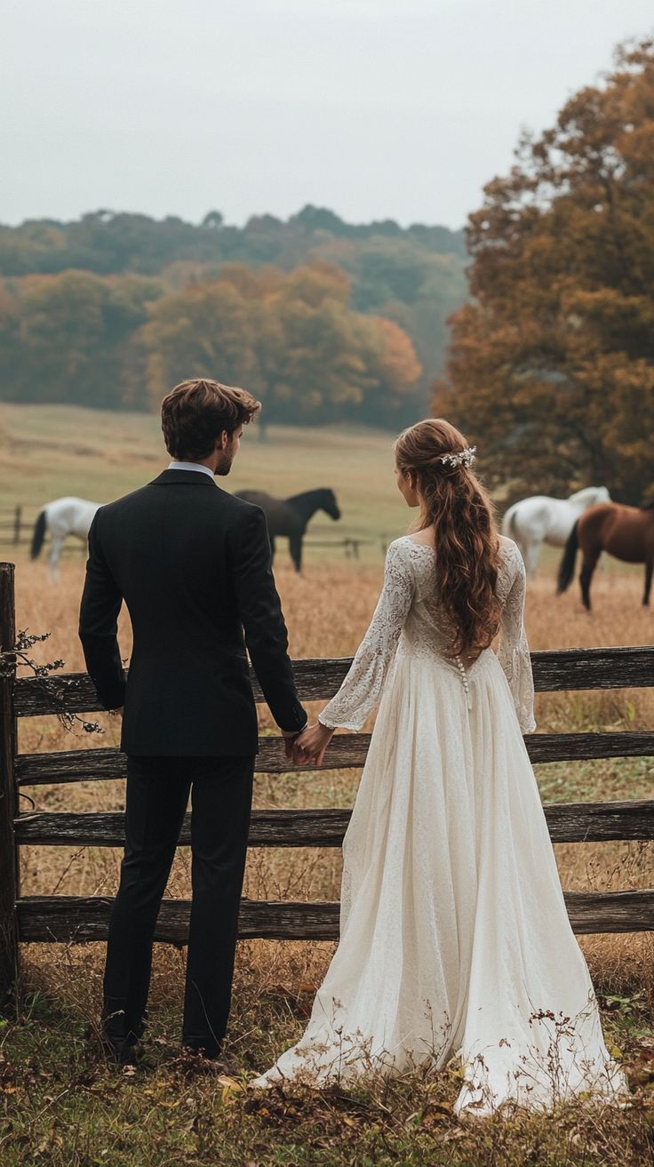 a bride and groom standing in front of a fence with horses grazing on the field behind them