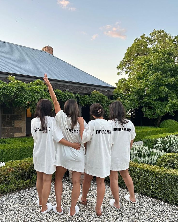 three girls in white shirts standing on gravel with their arms up and one girl pointing to the sky