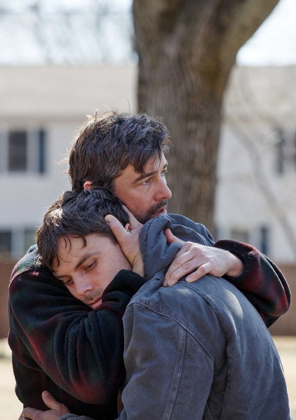 two men hugging each other in front of a tree and building with no leaves on it