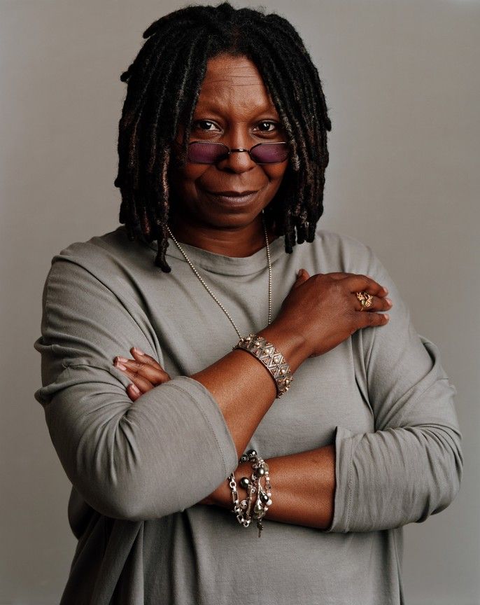 a woman with dreadlocks standing in front of a white wall wearing sunglasses and a gray shirt