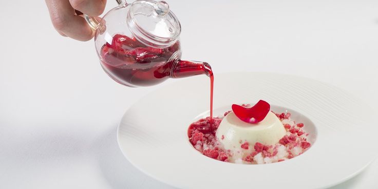 someone pouring sauce onto a dessert on a white plate with red crumbled toppings