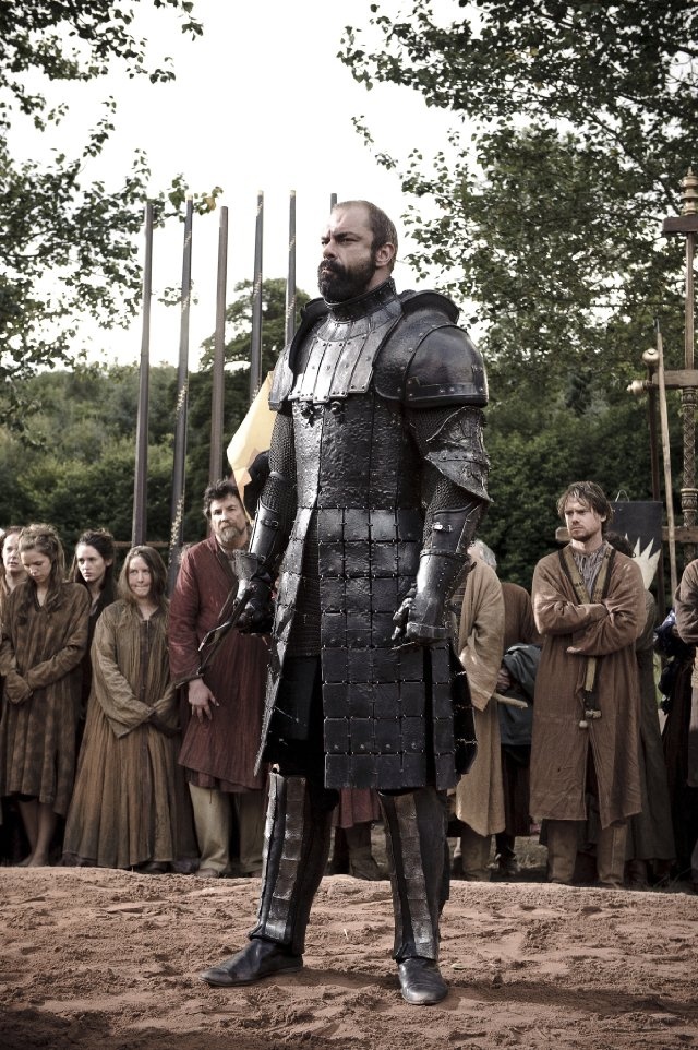 a man dressed in armor standing on top of a dirt field next to other people