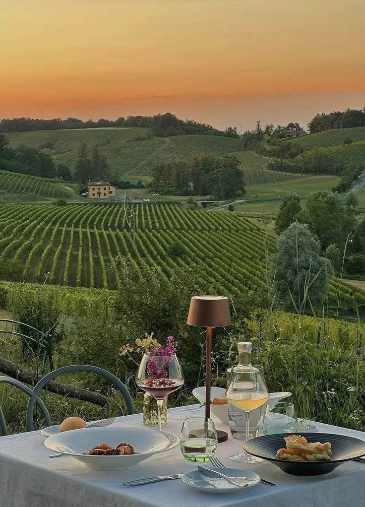 a table with food and wine on it in the middle of a vineyard field at sunset