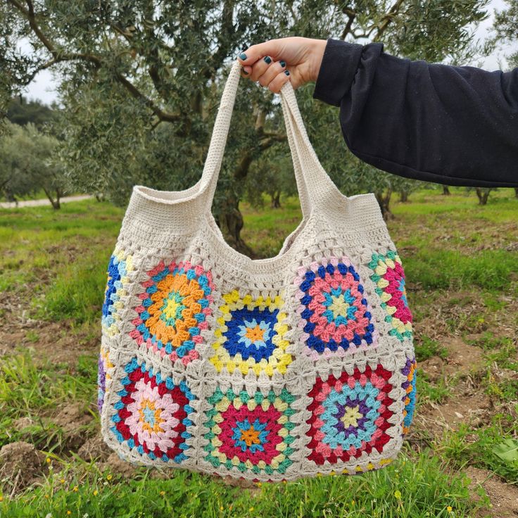 a hand holding a crocheted bag in the grass with an olive tree in the background