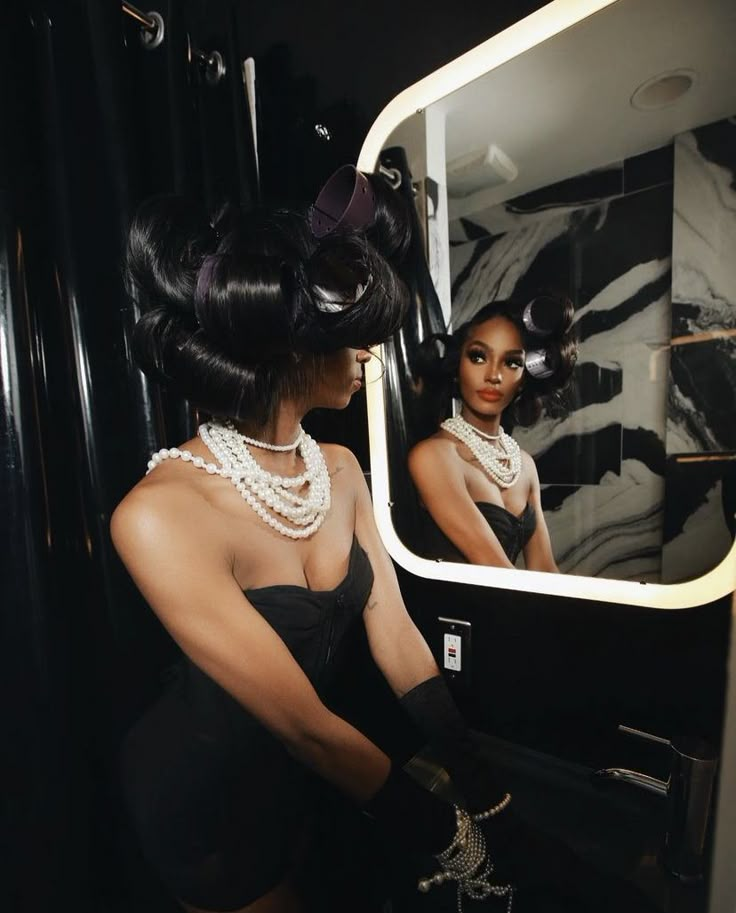 a woman in a black dress looking at herself in the mirror with pearls on her neck