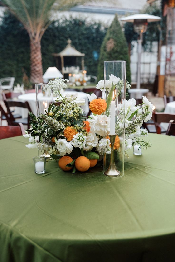 an arrangement of flowers and candles on top of a green table cloth covered round table