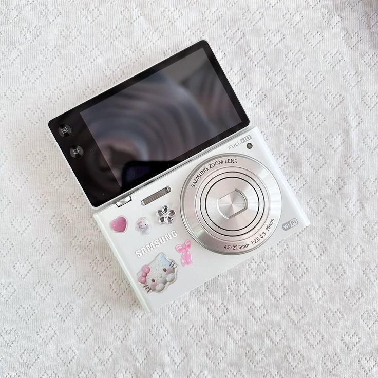 an electronic camera sitting on top of a white tablecloth covered bed sheet with cat stickers attached to the screen