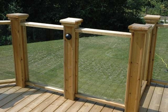 a wooden deck with glass railings in front of grass and trees on the other side