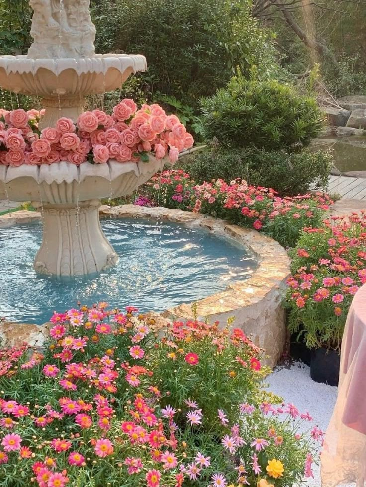 a fountain with pink flowers in it surrounded by greenery and other colorful flowerbedding