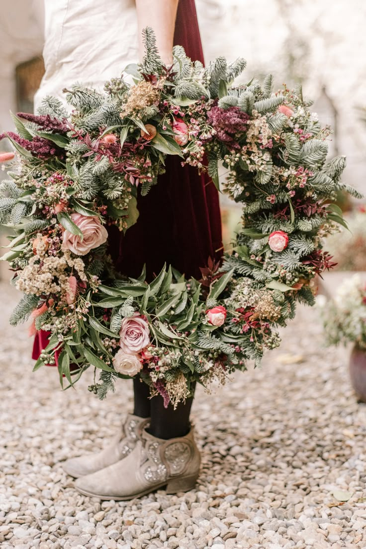 a woman is holding a wreath with flowers on it and boots in front of her