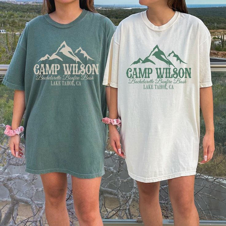 two girls standing next to each other in front of a fence and mountains with the words camp wilson on them