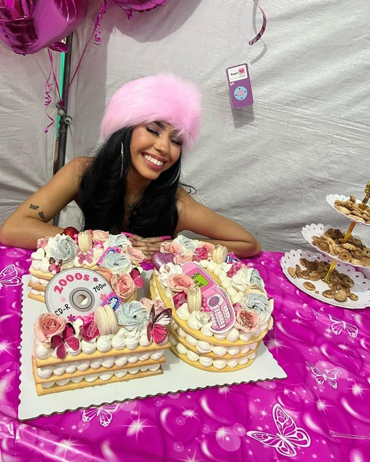 a woman sitting at a table with two large cakes on top of it and balloons in the background