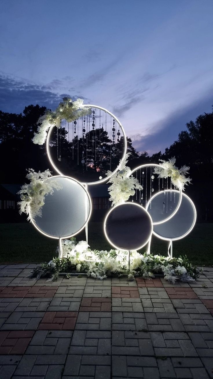 an outdoor display with circular mirrors and flowers on the ground at night, lit up by lights