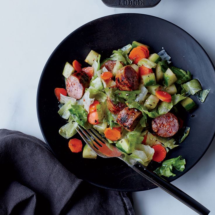 a black plate topped with salad next to a knife and fork on top of a white table