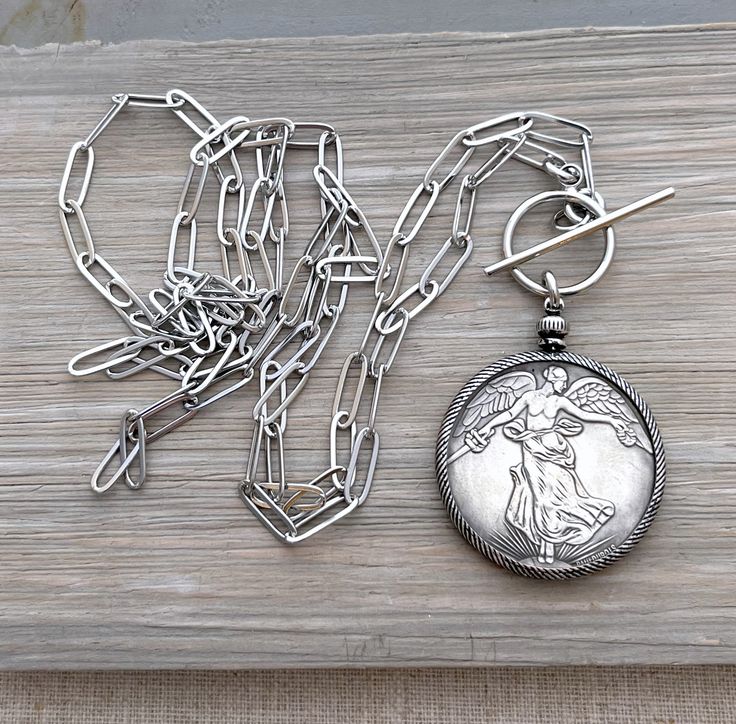 Please note - this item cannot be guaranteed to arrive for Christmas delivery. A beautiful replica antique French Victory coin cast in silver pewter is encircled in a silver plated brass coin holder with an 8mm brass ring bail. It hangs from a pewter silver paperclip style chain with a front closure oversized toggle clasp. Also available with a back closure in a lobster claw clasp. The coin with bezel measures 2 inches long X 1.5 inch wide.  Please see photo for image of reverse side of coin des Antique Silver Medallion Necklace With Locket, Heirloom Silver Necklace With Coin Pendant, Silver Medallion Necklace With Vintage Charm, Antique Engraved Medallion Coin Necklace, Antique Sterling Silver Medallion Coin Necklace, Vintage Silver Coin Necklace With Charm, Silver Vintage Charm Coin Necklace, Vintage Sterling Silver Medallion Necklace With Coin Pendant, Silver Vintage Coin Necklace With Charm