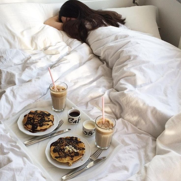 a woman laying in bed with two plates of food and drinks on the table next to her