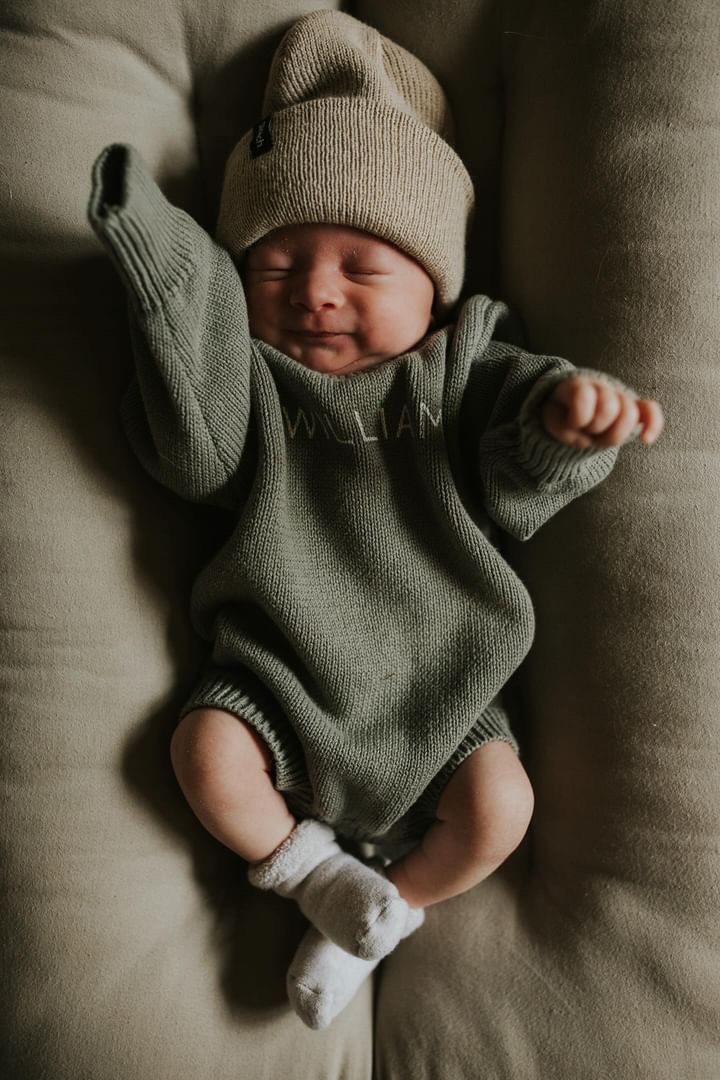 a baby wearing a hat and sweater laying on top of a couch next to a pillow