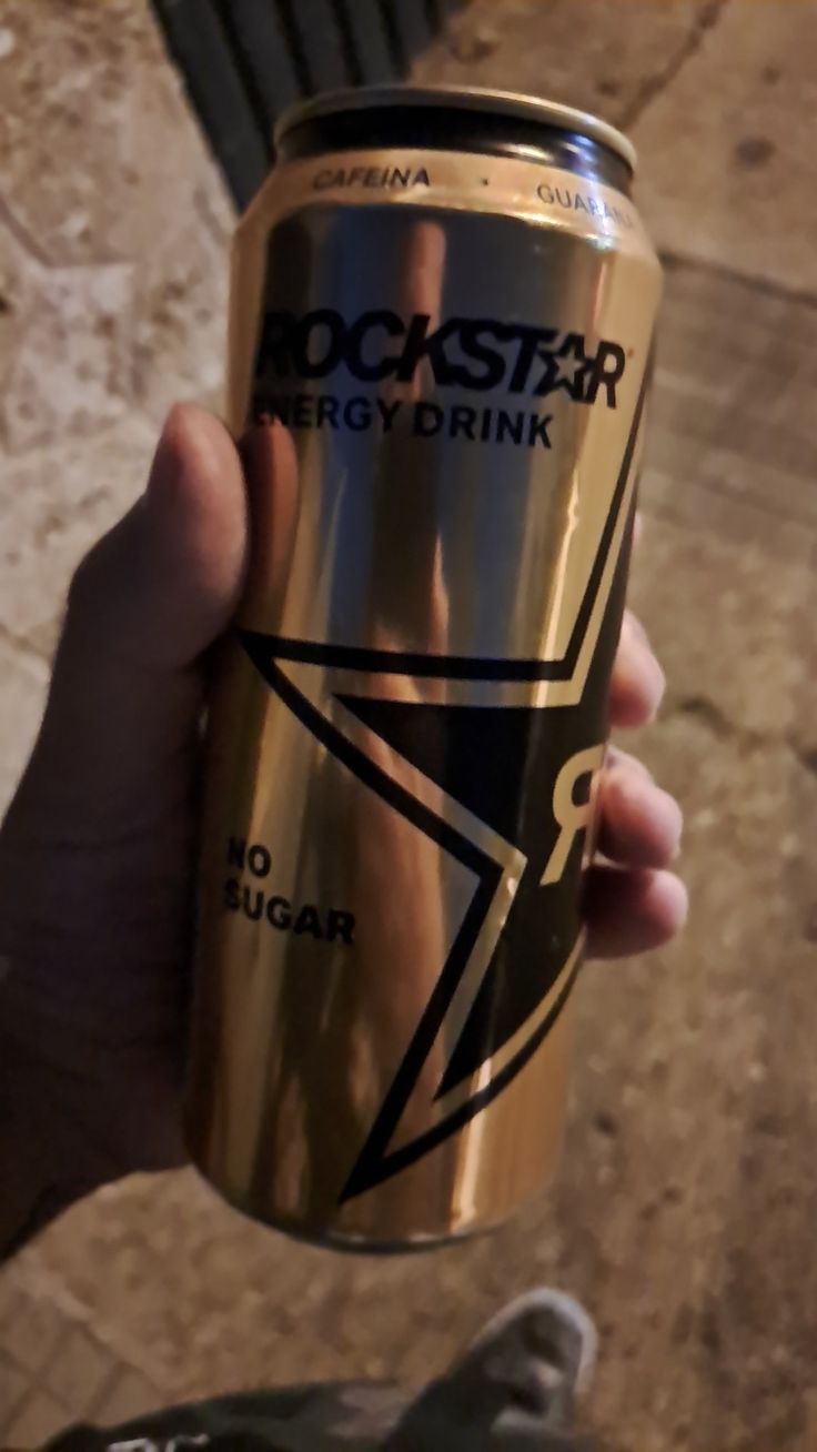 a person holding up a can of rock star energy drink in their left hand, on the ground