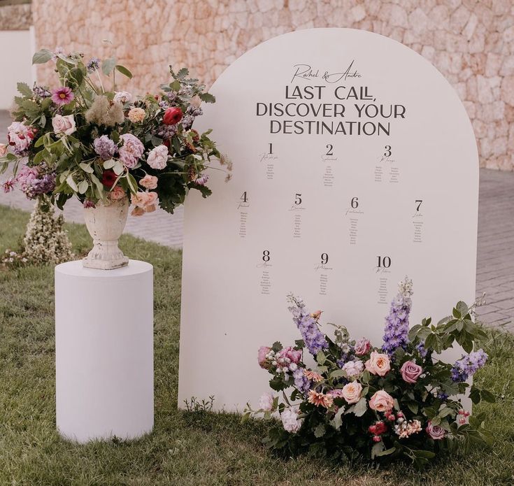 a large white sign sitting on top of a lush green field next to a vase filled with flowers